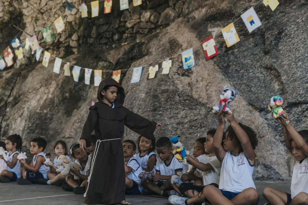 Junge in Brauner Robe geht an Kindern die auf dem Boden sitzen vorbei