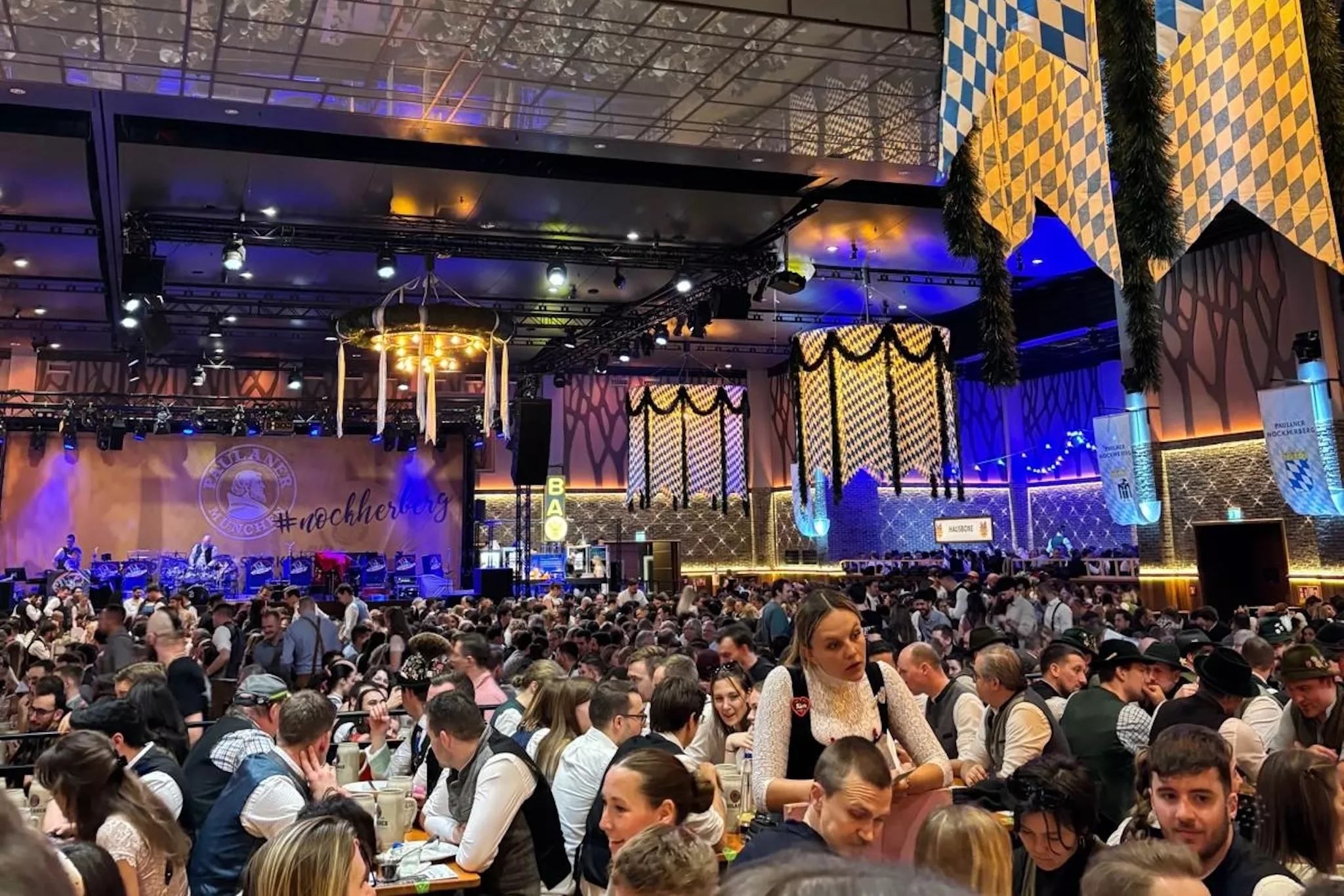 Viele Besucher des Paulaner am Nockherberg in München sitzen auf Bierbänken. Im Hintergrund sind Bayernfahnen zu sehen.