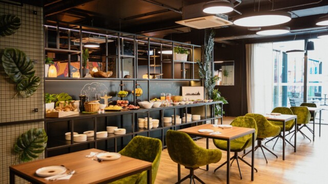 Holztische mit grünen Stühlen vor Frühstücksbuffet