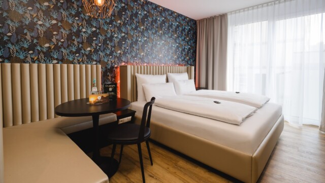 Großes Doppelbett mit weißem Laken. Daneben befindet sich ein Tisch, ein Stuhl und eine Sitzbank.