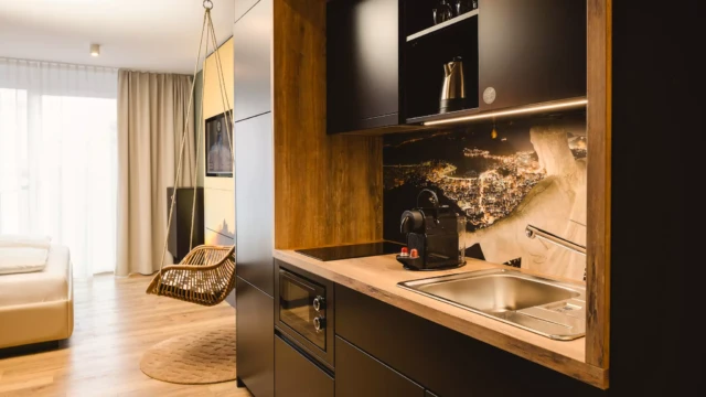 Küchenzeile in schwarz in Hotel mit Kaffemaschine, Herdplatte und Wasserkocher. Im Hintergrund befindet sich ein Hängestuhl, ein TV und ein großes Doppelbett.