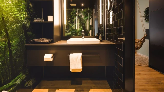 Modernes Hotelbad mit schwarzen Schränken und weißem Waschbecken. Über dem Waschbecken ist ein großer beleuchteter Spiegel.