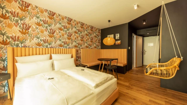 Großes Doppelbett mit weißem Laken. Dahinter bunte Tapete und Sitzecke.